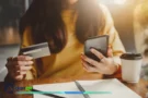 Mujer con tarjeta y celular revisa costos ocultos de su tarjeta de crédito.