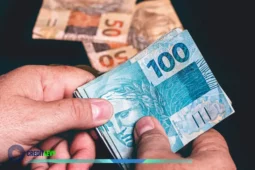 Pessoa segurando notas de 100 reais com cédulas ao fundo, simbolizando o auxílio emergencial e o apoio financeiro oferecido a cidadãos em situação de vulnerabilidade.