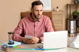Hombre revisando su estado de cuenta en el celular mientras trabaja frente a una laptop.